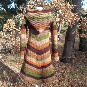 Puede incluir: Abrigo largo con capucha de ganchillo con un patrón de chevrón. El abrigo presenta una mezcla de colores tierra, incluyendo marrón, verde, naranja y beige. El abrigo se muestra en un maniquí blanco al aire libre.