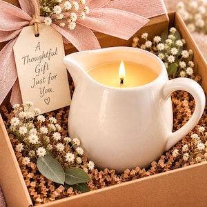 May include: A white ceramic pitcher candle with a lit wick, nestled in a brown gift box filled with crinkle paper and white baby's breath flowers. A pink ribbon and a beige tag with the text A Thoughtful Gift Just for You are attached.