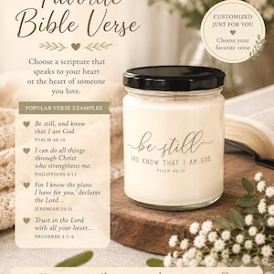 May include: A clear glass candle jar with a black lid, featuring the white text "be still AND KNOW THAT I AM GOD. PSALM 46:10" printed on the front. The candle is filled with white wax and sits on a dark wooden surface with small white flowers.