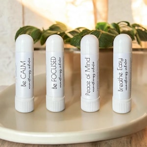 May include: Four white aromatherapy inhalers with the phrases "Be Calm", "Be Focused", "Peace of Mind", and "Breathe Easy" printed on them. The inhalers are arranged on a light-colored, round dish, with green foliage in the background.