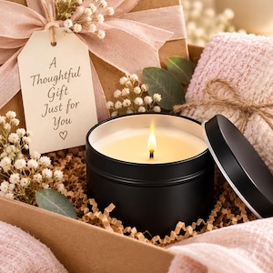 May include: A gift box containing a black metal candle tin with a lit candle, a pink textured fabric, and a beige gift tag with the text A Thoughtful Gift Just for You and a heart symbol, decorated with a pink ribbon and small white flowers.
