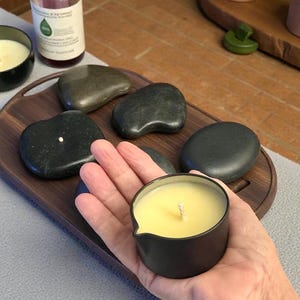 May include: A small, round, matte black candle container with a pouring spout, filled with pale yellow wax and a central wick. It is held above a dark wooden tray containing several smooth, dark grey river stones and a small glass bottle of oil.