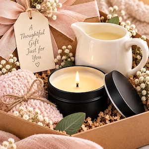 May include: A gift box containing a lit candle in a black tin with a matching lid, a white ceramic creamer, a pink textured fabric item, and a tag with the text A Thoughtful Gift Just for You with a heart symbol, all accented with baby's breath.