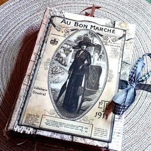 May include: A rectangular journal featuring a vintage-style cover with the text Au Bon Marché and Ete 1911. The cover displays a black and white illustration of a person in a long dress and hat, accented with lace trim and a dark grey ribbon bow.