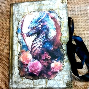 Peut inclure: Un journal fait à la main avec une illustration de dragon. La couverture présente une image détaillée d'un dragon dans des tons de bleu, de violet et de rouge, entouré de fleurs roses. Le journal a un aspect vieilli et texturé avec des accents dorés et un ruban noir.