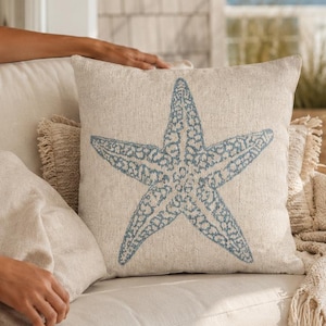 May include: A square throw cushion featuring a light beige linen-textured cover with a prominent blue starfish graphic printed in the centre. The starfish design displays a detailed, textured pattern across its surface, contrasting with the neutral fabric.