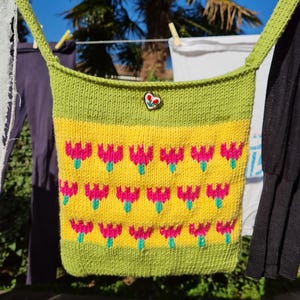 Può includere: Borsa a tracolla lavorata a maglia a mano con un vivace design floreale. La borsa presenta un pannello centrale giallo ornato di fiori rosa e verdi a forma di tulipano. La parte superiore e inferiore sono verdi, e anche la tracolla è verde. È attaccata una spilla a forma di cuore.