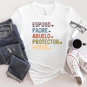 May include: White t-shirt with the words "ESPOSO, PADRE, ABUELO, PROTECTOR, HEROE" in colourful text. The shirt is paired with blue jeans, white trainers, a black coffee mug, and black glasses.