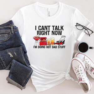 Può includere: T-shirt bianco con la scritta "I CAN'T TALK RIGHT NOW I'M DOING HOT DAD STUFF" e illustrazioni di una griglia, una cassetta degli attrezzi, boccali di birra e un tosaerba. La maglietta è abbinata a jeans blu, scarpe da ginnastica bianche e una tazza da caffè nera.
