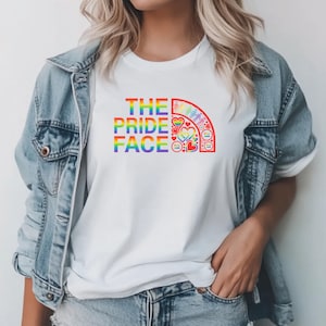 Puede incluir: Camiseta blanca con el texto "THE PRIDE FACE" en colores arcoíris. El diseño incluye un arco iris con corazones y otros símbolos. La persona lleva una chaqueta vaquera.