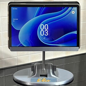 Può includere: Un supporto per tablet grigio con base girevole. Il tablet mostra uno schermo blu con l'ora 00:03. Il supporto è su una superficie nera.
