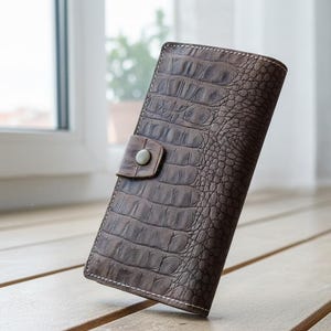 Puede incluir: Una cartera de cuero marrón con textura de piel de cocodrilo. La cartera tiene un cierre de botón a presión y costuras blancas en los bordes. La cartera mide aproximadamente 18 cm de alto y 10 cm de ancho.