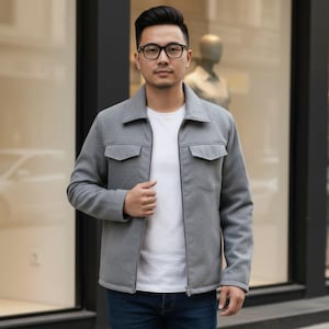 May include: A gray jacket with a zipper closure, two chest pockets, and a collared neckline. The jacket is worn over a white t-shirt. The person in the image is wearing glasses.