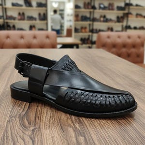 May include: Black leather sandal with woven detailing on the vamp and heel. The sandal features a buckle closure and a low, flat sole. The shoe is displayed on a wooden surface.