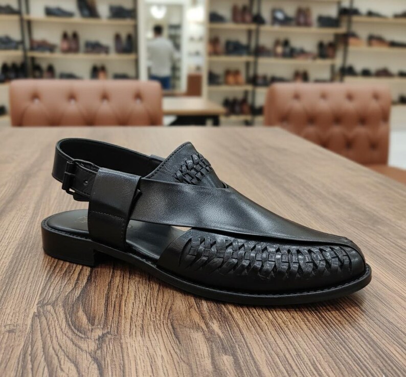 May include: Black leather sandal with woven detailing on the vamp and heel. The sandal features a buckle closure and a low, flat sole. The shoe is displayed on a wooden surface.