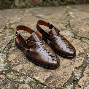 May include: A pair of dark brown leather huarache sandals featuring a woven upper design, a closed toe, and an adjustable ankle strap with a gold-toned metal buckle. The sandals have a smooth finish and a dark sole, showcasing a classic construction.