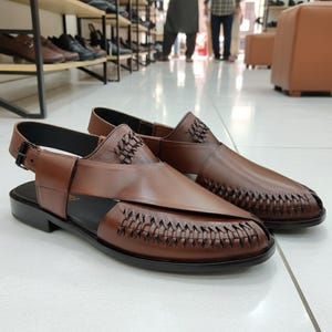 May include: A pair of brown leather sandals with woven detailing on the toe and sides. The sandals have a slingback strap with a buckle closure and a black sole. The shoes are displayed on a white tiled floor.