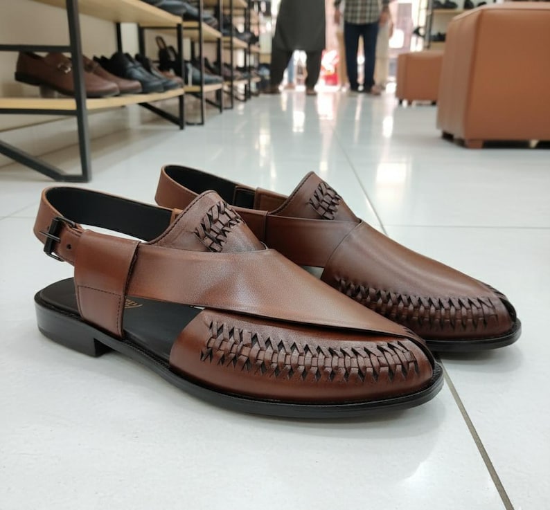 May include: A pair of brown leather sandals with woven detailing on the toe and sides. The sandals have a slingback strap with a buckle closure and a black sole. The shoes are displayed on a white tiled floor.