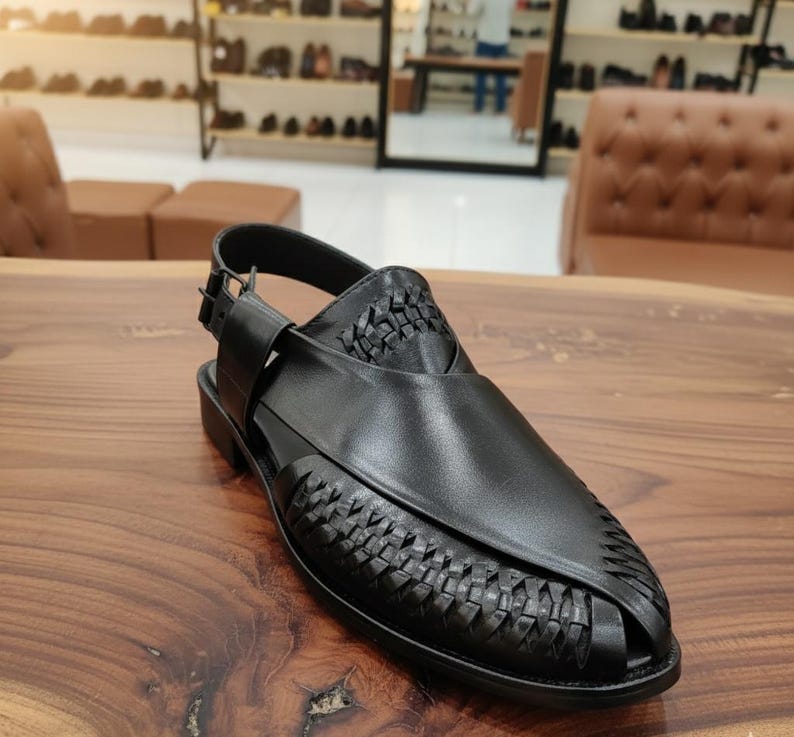 May include: Black leather sandal with woven detailing on the vamp and heel. The sandal features an adjustable ankle strap with a buckle closure. The shoe has a low heel and is designed for comfort and style. The sandal is displayed on a wooden surface.