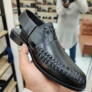 May include: Black leather Peshawari sandal with woven detailing on the vamp. The sandal features a strap around the ankle and a low heel. The shoe is held by a person in a store setting, with other shoes visible in the background.