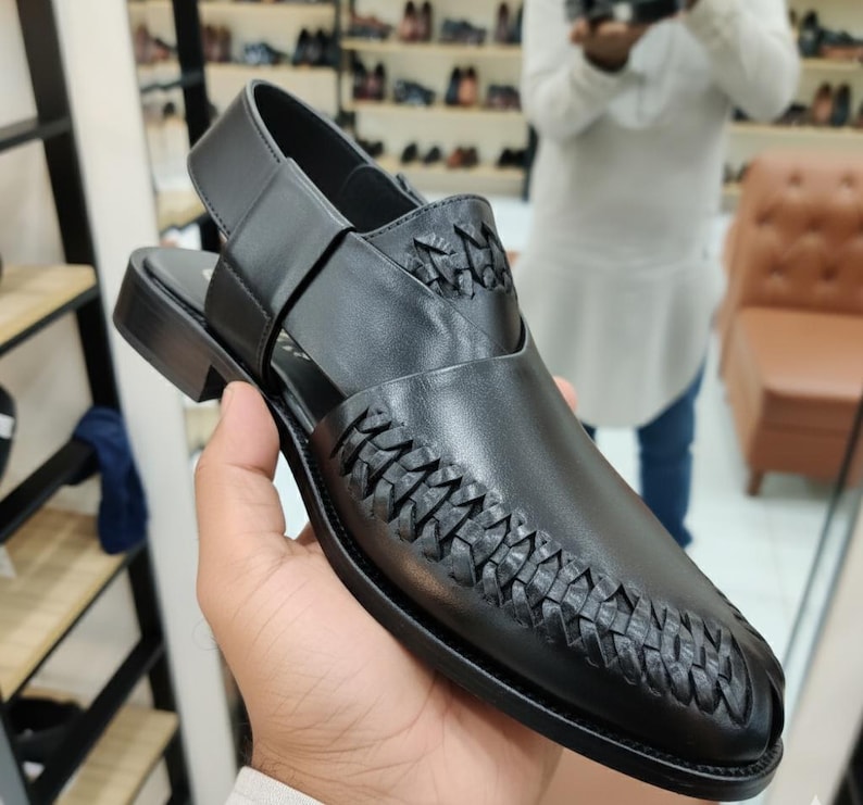 May include: Black leather Peshawari sandal with woven detailing on the vamp. The sandal features a strap around the ankle and a low heel. The shoe is held by a person in a store setting, with other shoes visible in the background.