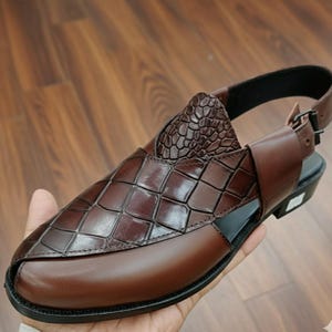 May include: Brown leather sandal with a geometric pattern on the upper and a textured heel. The sandal has a buckle closure and a black sole. The shoe is held in a person's hand.