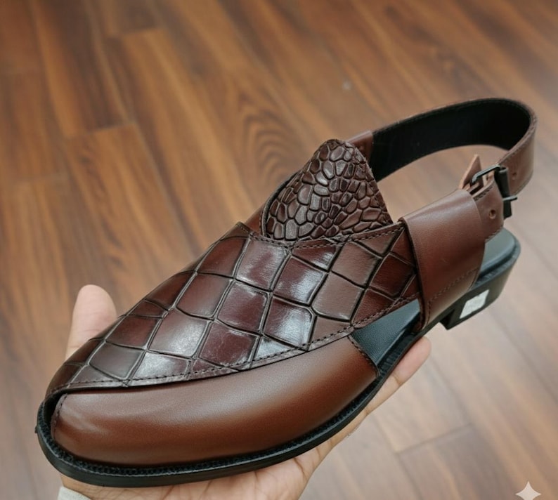 May include: Brown leather sandal with a geometric pattern on the upper and a textured heel. The sandal has a buckle closure and a black sole. The shoe is held in a person's hand.