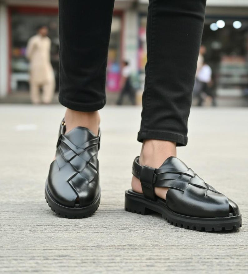 May include: Black leather sandals with a woven design on the upper and a thick, lugged sole. The sandals have an adjustable ankle strap and are worn with black pants. The background is blurred, suggesting an outdoor setting.