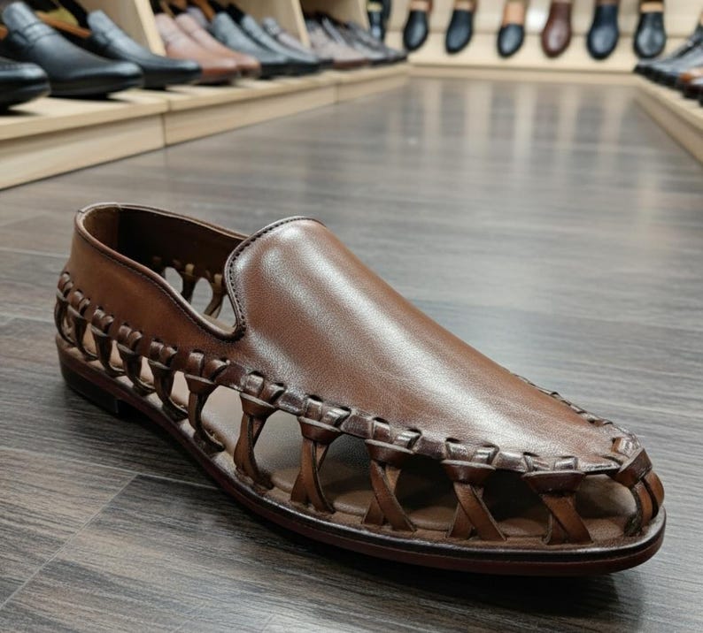 K&ouml;nnte beinhalten: Brauner Leder-Slipper mit gewebten Seitendetails. Der Schuh hat ein glattes, poliertes Obermaterial und eine flache Sohle. Weitere Schuhe sind im Hintergrund zu sehen.