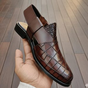 May include: A brown leather sandal with a woven pattern on the vamp and a textured accent. The sandal has a slingback strap and a small heel. The shoe is held in a hand.