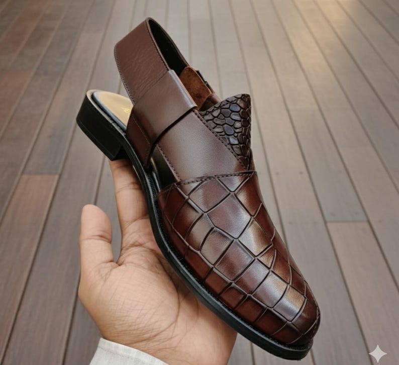 May include: A brown leather sandal with a woven pattern on the vamp and a textured accent. The sandal has a slingback strap and a small heel. The shoe is held in a hand.