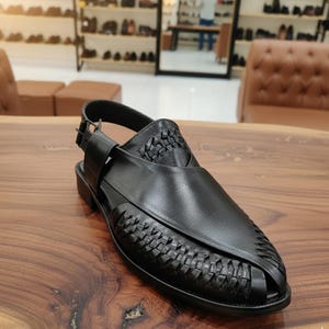 May include: Black leather sandal with woven detailing on the vamp and heel. The sandal features an adjustable ankle strap with a buckle closure. The shoe is set on a low, black heel and is displayed on a wooden surface.