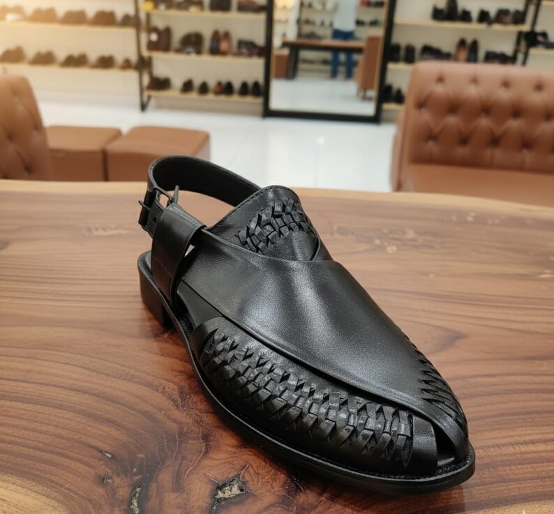May include: Black leather sandal with woven detailing on the vamp and heel. The sandal features an adjustable ankle strap with a buckle closure. The shoe is set on a low, black heel and is displayed on a wooden surface.