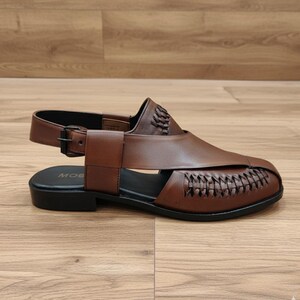 May include: Brown leather sandal with a woven design on the front. The sandal has a strap around the ankle with a buckle closure. The sole is black. The word "MOG" is visible on the insole.