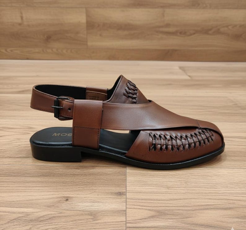 May include: Brown leather sandal with a woven design on the front. The sandal has a strap around the ankle with a buckle closure. The sole is black. The word "MOG" is visible on the insole.