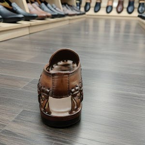 K&ouml;nnte beinhalten: Eine braune Ledersandale mit gewebten Details. Der offene Schuh hat eine dunkle Sohle und wird auf einem Holzboden pr&auml;sentiert. Weitere Schuhe sind im Hintergrund auf Regalen zu sehen.
