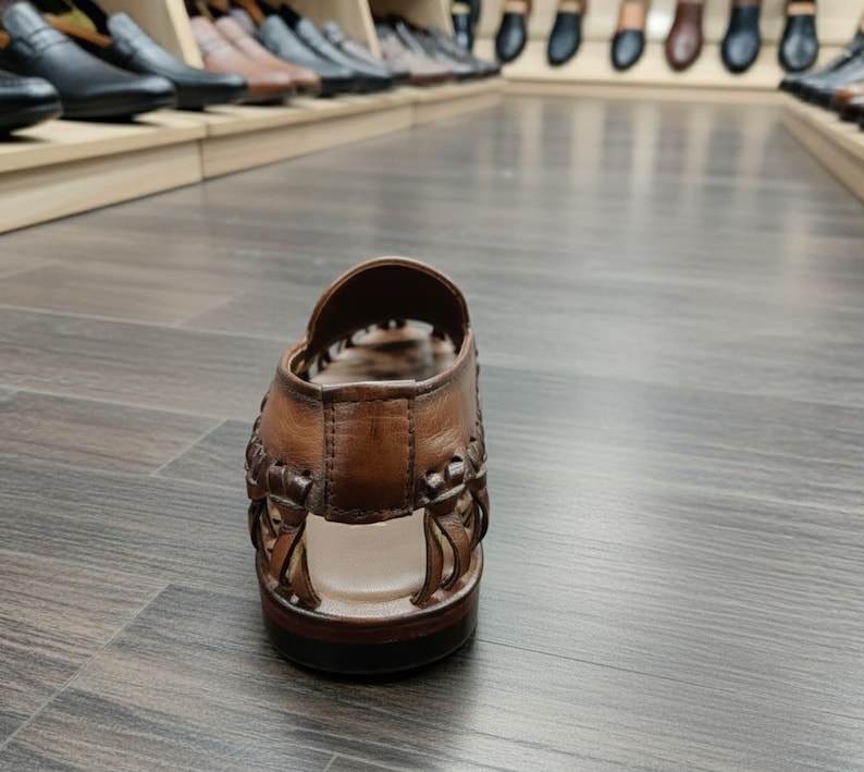 K&ouml;nnte beinhalten: Eine braune Ledersandale mit gewebten Details. Der offene Schuh hat eine dunkle Sohle und wird auf einem Holzboden pr&auml;sentiert. Weitere Schuhe sind im Hintergrund auf Regalen zu sehen.