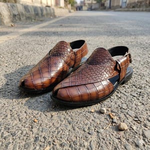 May include: A pair of brown leather sandals with a crocodile skin texture. The sandals have a strap around the ankle and a buckle closure. The shoes are on a black sole.