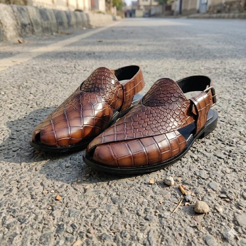May include: A pair of brown leather sandals with a crocodile skin texture. The sandals have a strap around the ankle and a buckle closure. The shoes are on a black sole.