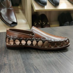 K&ouml;nnte beinhalten: Brauner Leder-Slipper mit gewebtem Design an den Seiten. Der Schuh hat eine runde Spitze und einen niedrigen Absatz. Weitere Schuhe sind auf Regalen im Hintergrund zu sehen.