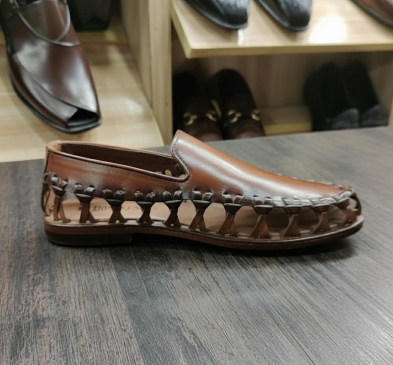 K&ouml;nnte beinhalten: Brauner Leder-Slipper mit gewebtem Design an den Seiten. Der Schuh hat eine runde Spitze und einen niedrigen Absatz. Weitere Schuhe sind auf Regalen im Hintergrund zu sehen.