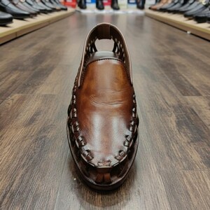 K&ouml;nnte beinhalten: Nahaufnahme eines braunen Lederschuhs mit gewebten Details an Zehen und Seiten. Der Schuh hat eine runde Spitze und ein Ausschnittdesign am Obermaterial. Der Schuh wird auf einem Holzboden pr&auml;sentiert, im Hintergrund sind weitere Schuhe zu sehen.