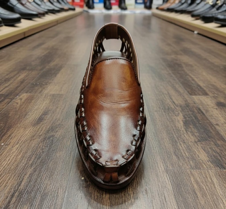 K&ouml;nnte beinhalten: Nahaufnahme eines braunen Lederschuhs mit gewebten Details an Zehen und Seiten. Der Schuh hat eine runde Spitze und ein Ausschnittdesign am Obermaterial. Der Schuh wird auf einem Holzboden pr&auml;sentiert, im Hintergrund sind weitere Schuhe zu sehen.