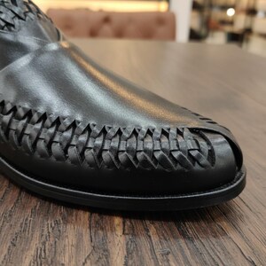 May include: Close-up of a black leather shoe with a woven design on the toe and upper. The shoe is set on a wooden surface, showcasing the texture and detail of the leather and the craftsmanship of the woven pattern.
