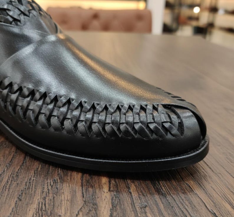 May include: Close-up of a black leather shoe with a woven design on the toe and upper. The shoe is set on a wooden surface, showcasing the texture and detail of the leather and the craftsmanship of the woven pattern.