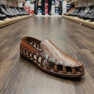 K&ouml;nnte beinhalten: Ein brauner Leder-Slipper mit geflochtenem Design an den Seiten. Der Schuh wird auf einem Holzboden pr&auml;sentiert, mit anderen Schuhen im Hintergrund. Der Schuh hat eine runde Spitze und ein glattes Obermaterial.