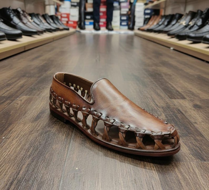 K&ouml;nnte beinhalten: Ein brauner Leder-Slipper mit geflochtenem Design an den Seiten. Der Schuh wird auf einem Holzboden pr&auml;sentiert, mit anderen Schuhen im Hintergrund. Der Schuh hat eine runde Spitze und ein glattes Obermaterial.
