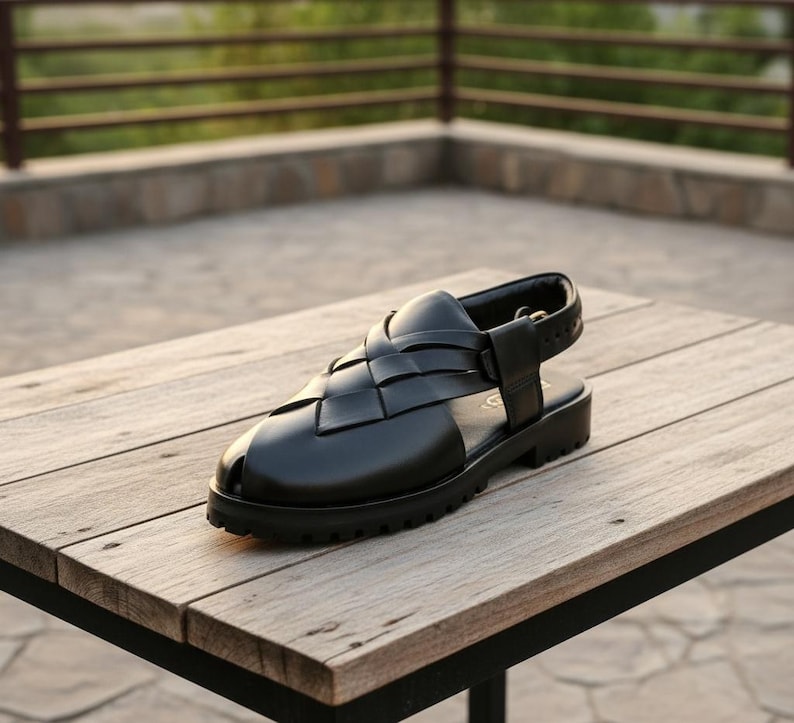 May include: Black leather sandal with a woven vamp design. The sandal features a thick, lug sole and an adjustable ankle strap. The shoe is displayed on a wooden table, showcasing its construction and style.