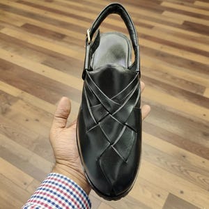 May include: Black leather sandal with a woven design on the upper. The sandal has a slingback strap with a buckle closure and a rounded toe. The shoe is held in a person's hand, showcasing the design and craftsmanship.