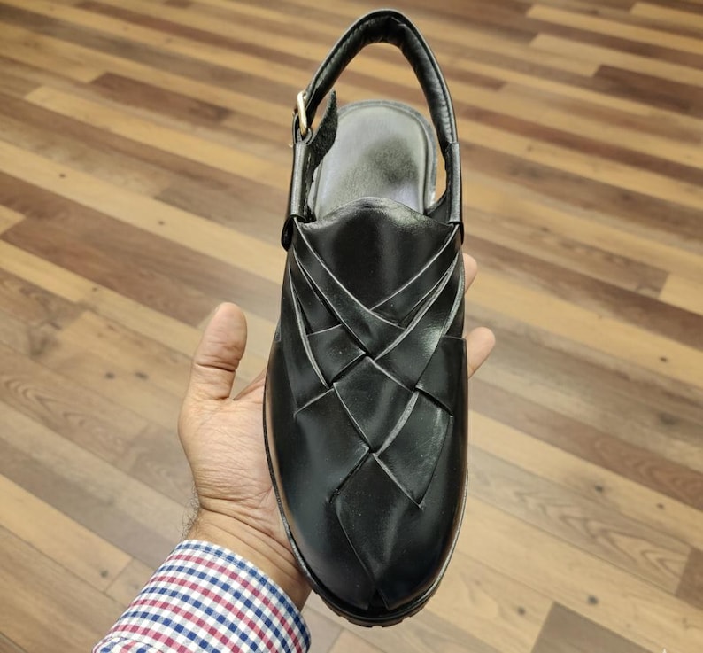 May include: Black leather sandal with a woven design on the upper. The sandal has a slingback strap with a buckle closure and a rounded toe. The shoe is held in a person's hand, showcasing the design and craftsmanship.